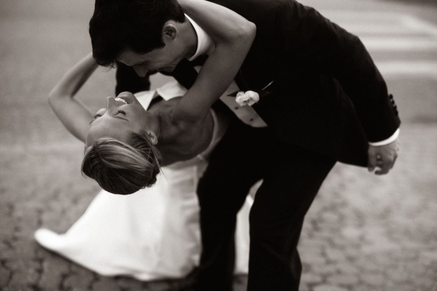 Bride in white strapless gown holding bouquet of white roses stands beside groom in black tuxedo under flowing veil.
