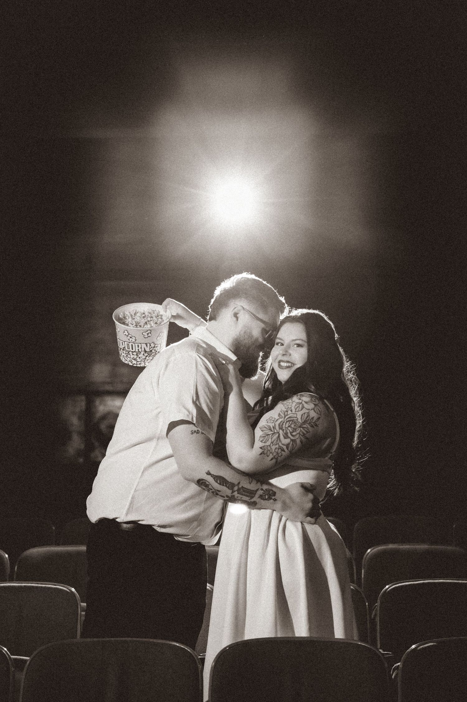 Black and white photo of a couple embracing and smiling in a theater, with a bright light glowing behind them.