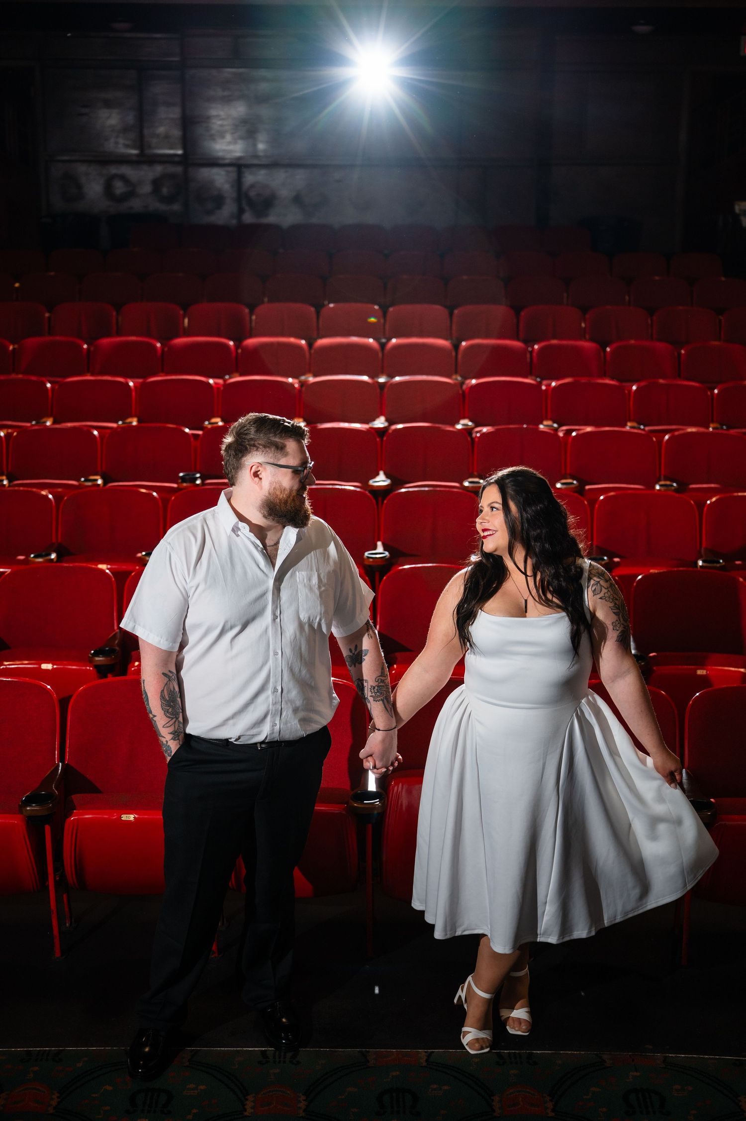 Couple holding hands in white outfits among red theater seats, smiling at each other under a spotlight.