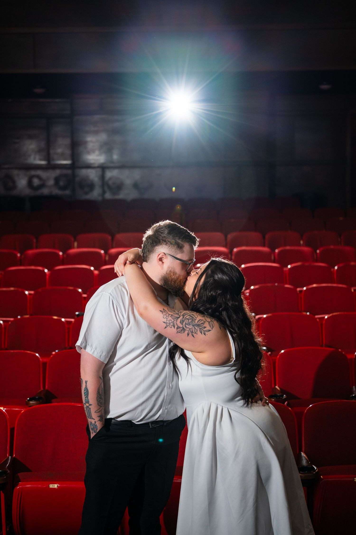 Couple sharing a romantic kiss in an empty red-seated theater with a bright spotlight glowing behind them.