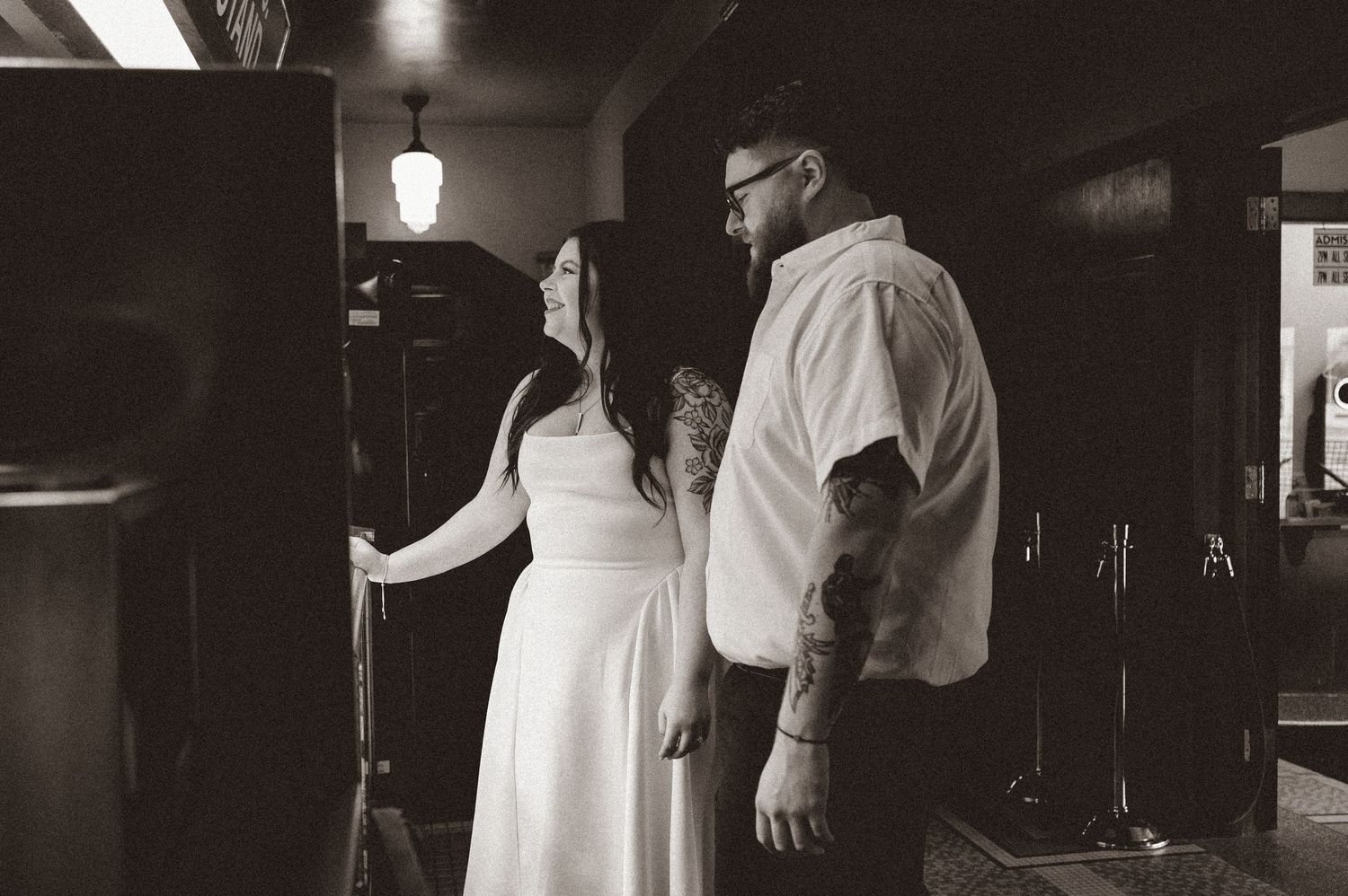Black and white photo of a couple in formal wear standing together indoors, man with tattoos wearing white shirt.