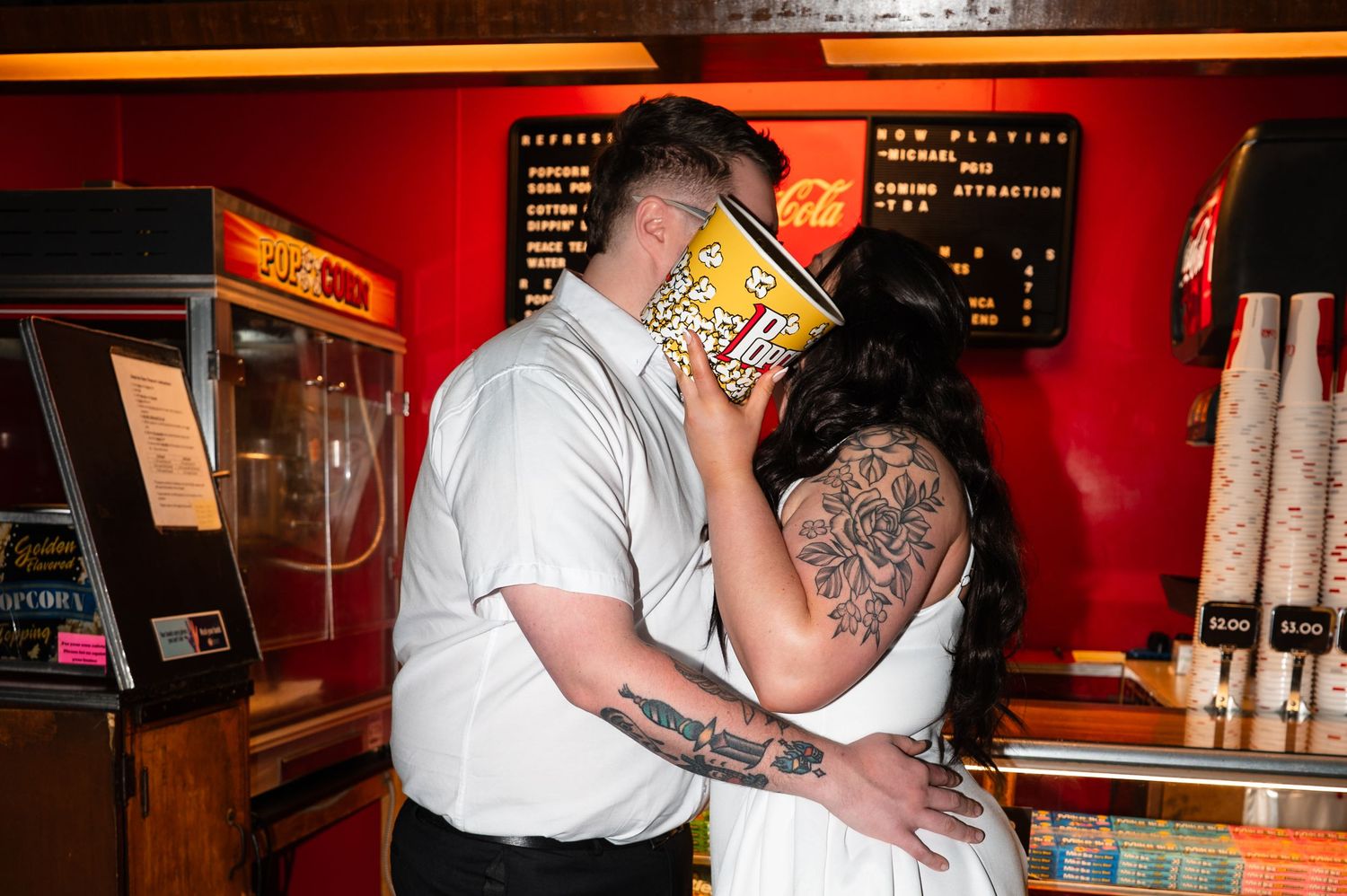 Couple sharing a kiss behind a large popcorn box at a movie theater concession stand.