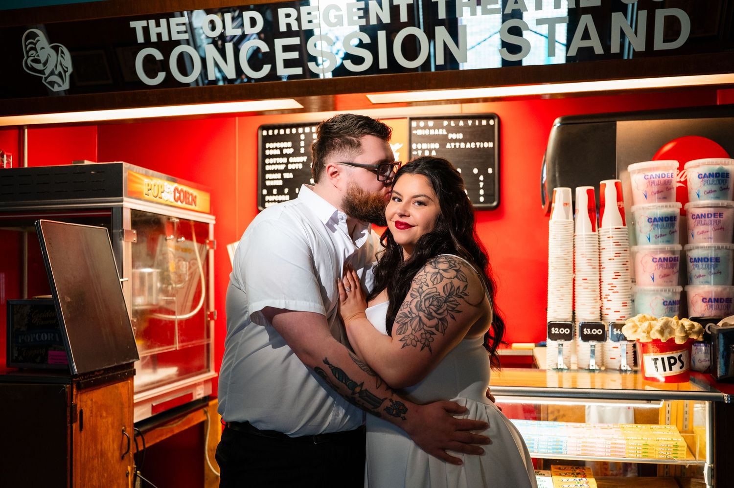 Couple embracing romantically at The Old Regent concessions stand, woman with tattoos in white dress, man in light shirt.