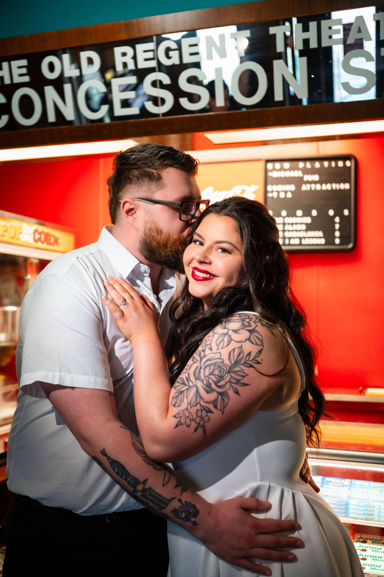 A tattooed couple embraces romantically at a vintage movie theater concession stand, the woman in white with red lips smiling.