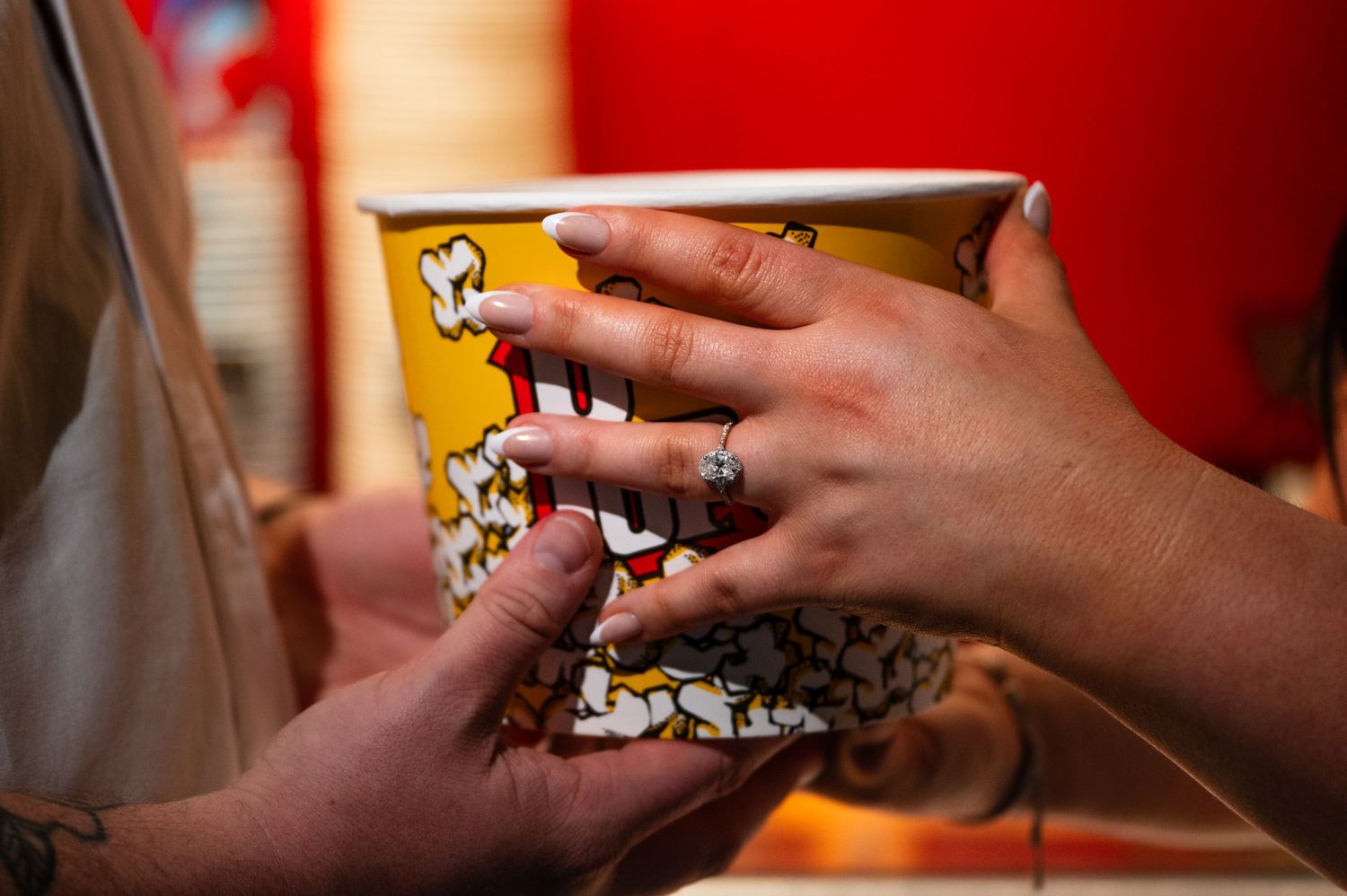 Two hands reaching into a large popcorn bucket at a movie theater, one hand wearing a diamond ring.