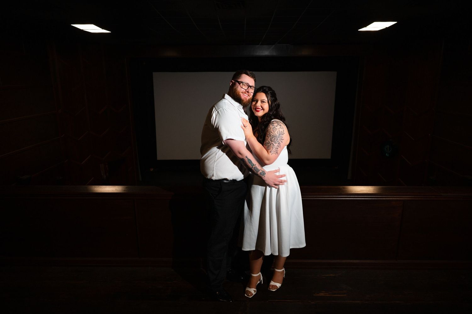 Couple embracing in front of a cinema screen, woman with tattoos in white dress, man in white shirt and black pants.