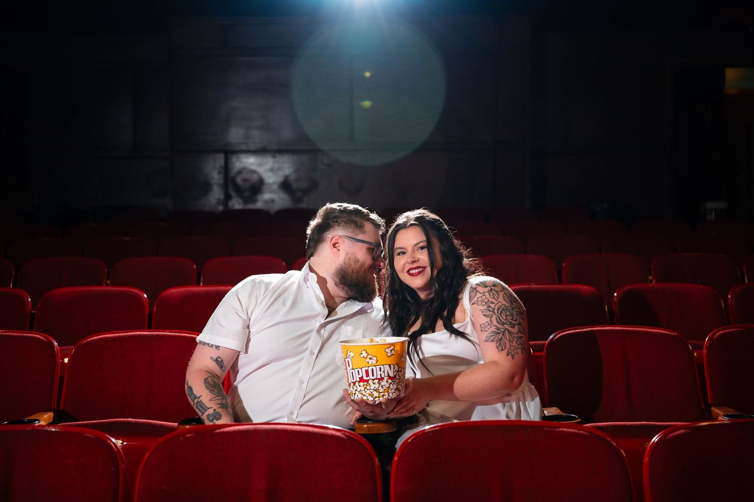 Couple sharing popcorn and a romantic moment together in a red-seated movie theater.