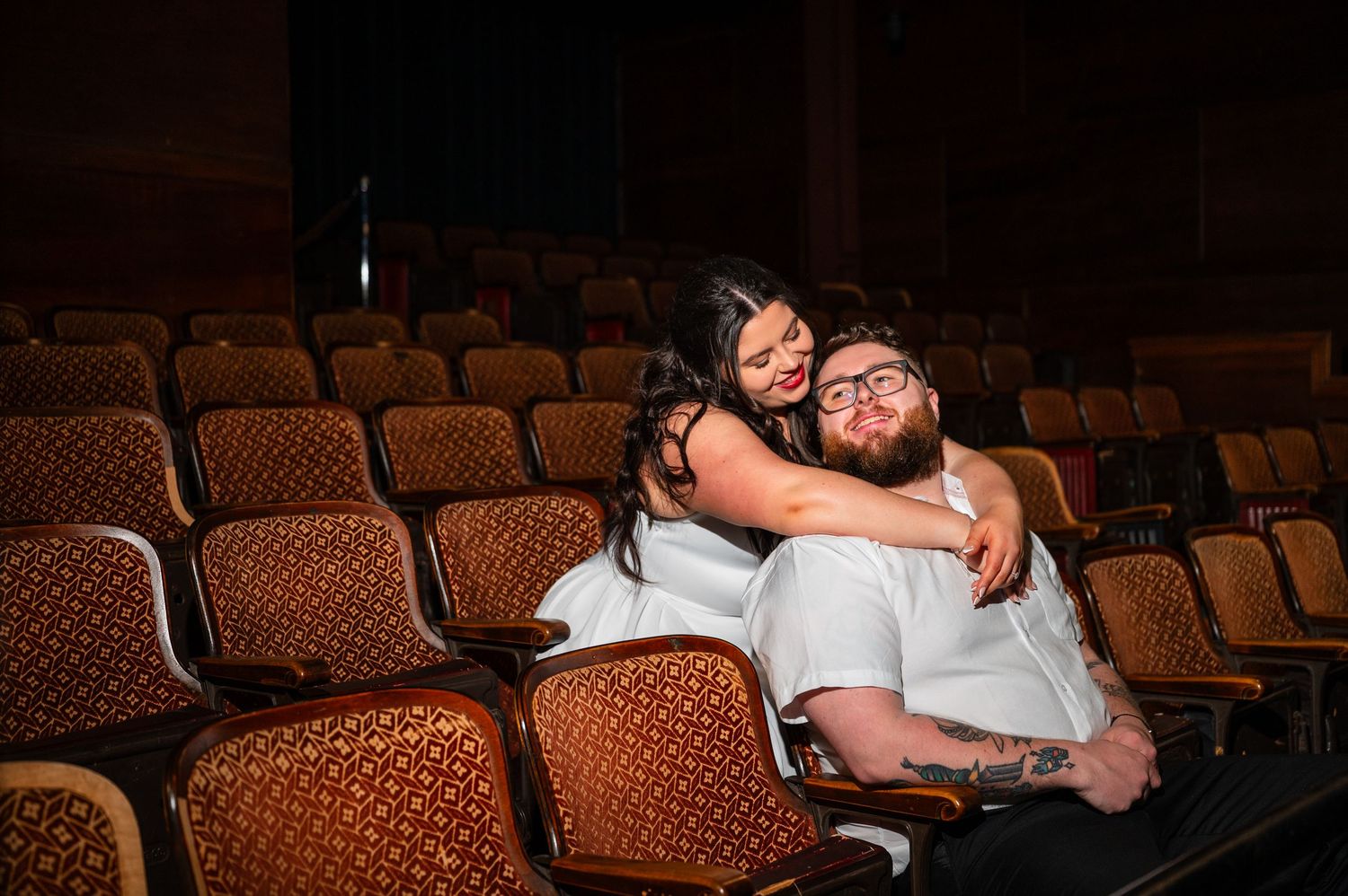 Couple embracing and smiling in vintage theater seats, woman hugging bearded man from behind in empty auditorium.