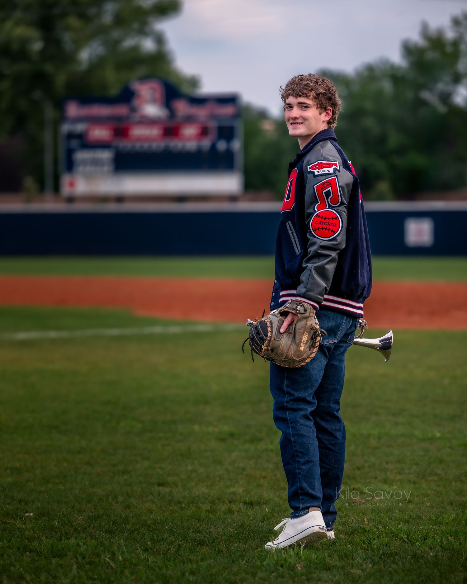 Teen boy in varsity jacket holding trumpet leans against brick wall outdoors at dusk for senior portrait photo.