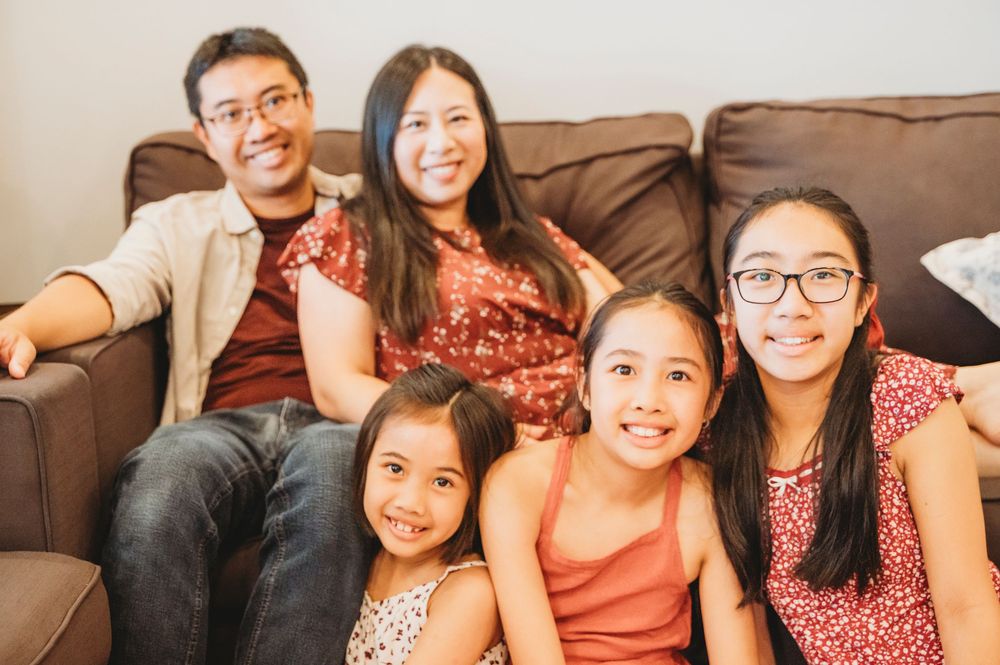 Mom and Dad sit on their couch in their home while their three daughters sit in front of them smiling.