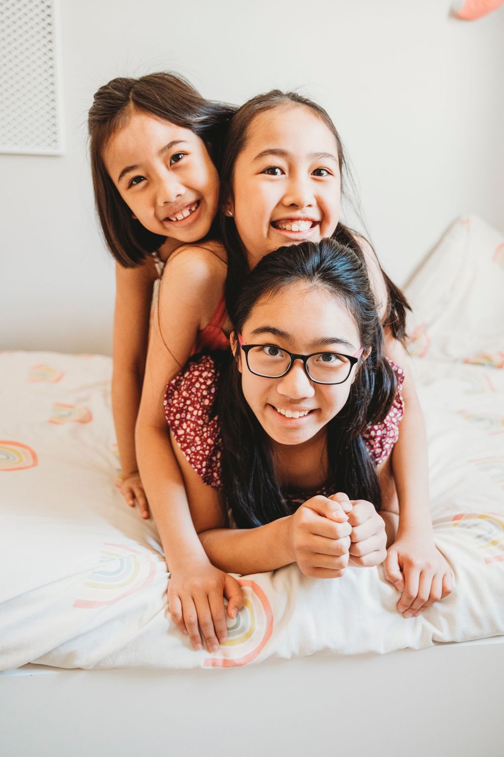 Three sisters lay stacked on one another on a white bed playing and smiling