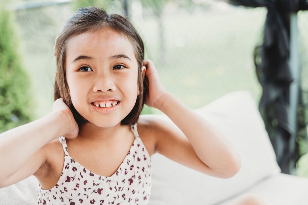 A little girl sits outside on a couch smiling while she tucks her hair between her ears