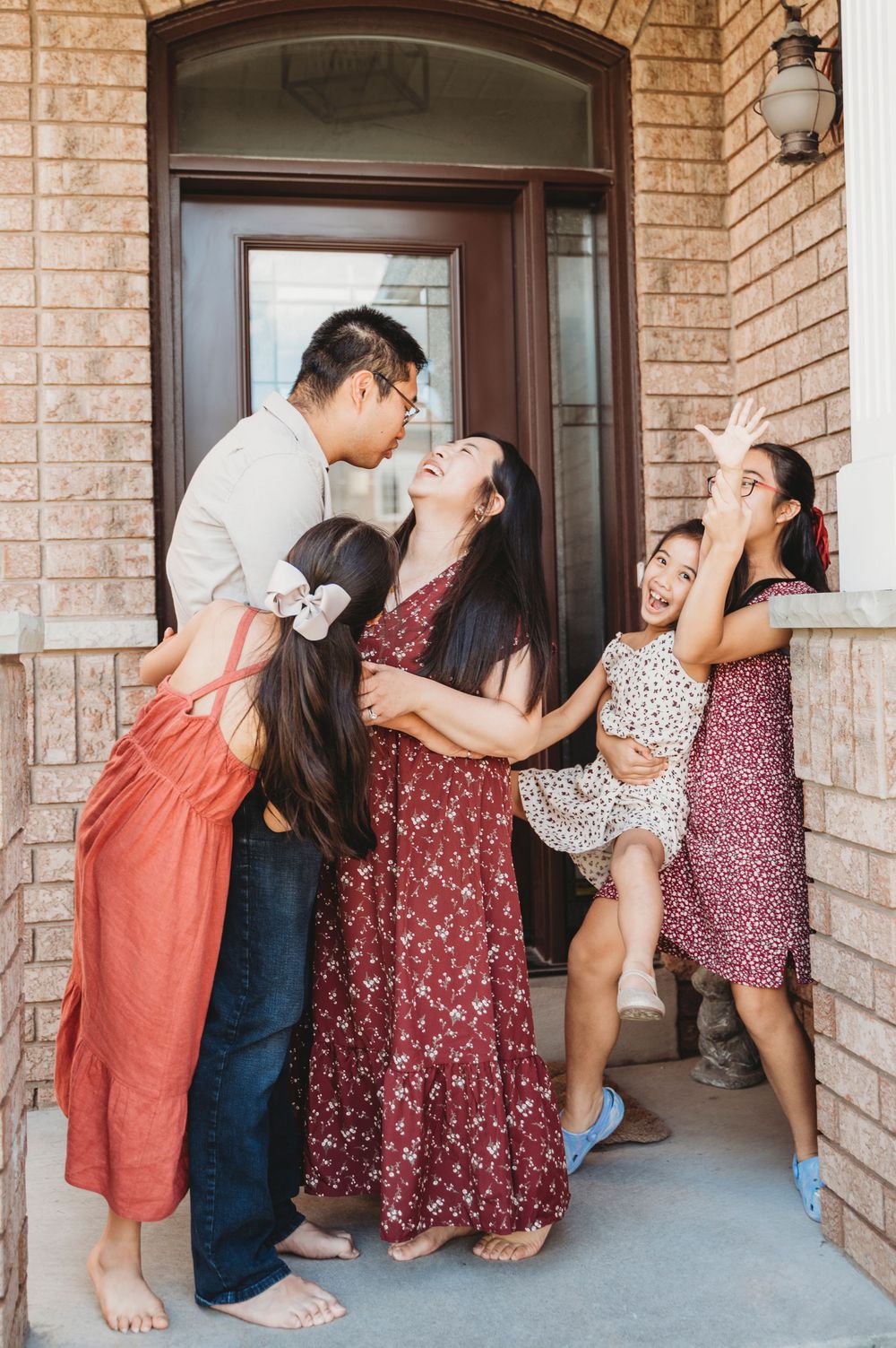 Mom and Dad embrace on their front porch while their three daughters make them laugh and play around them