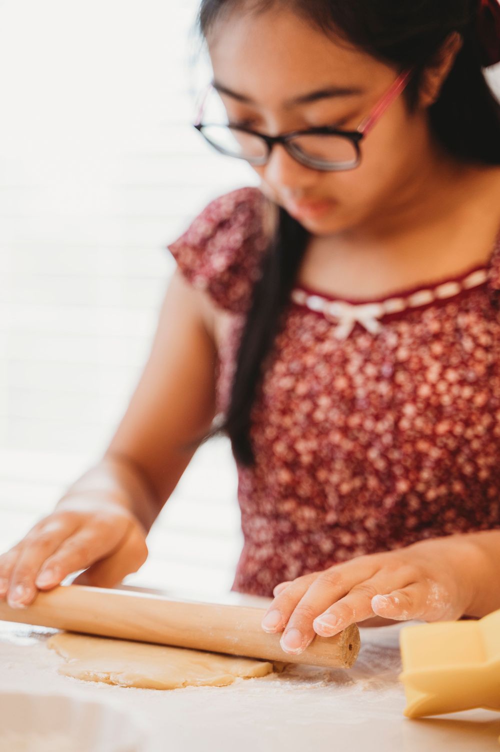 A little girl looks down and concentrates while rolling out cookie dough in her white kitchen