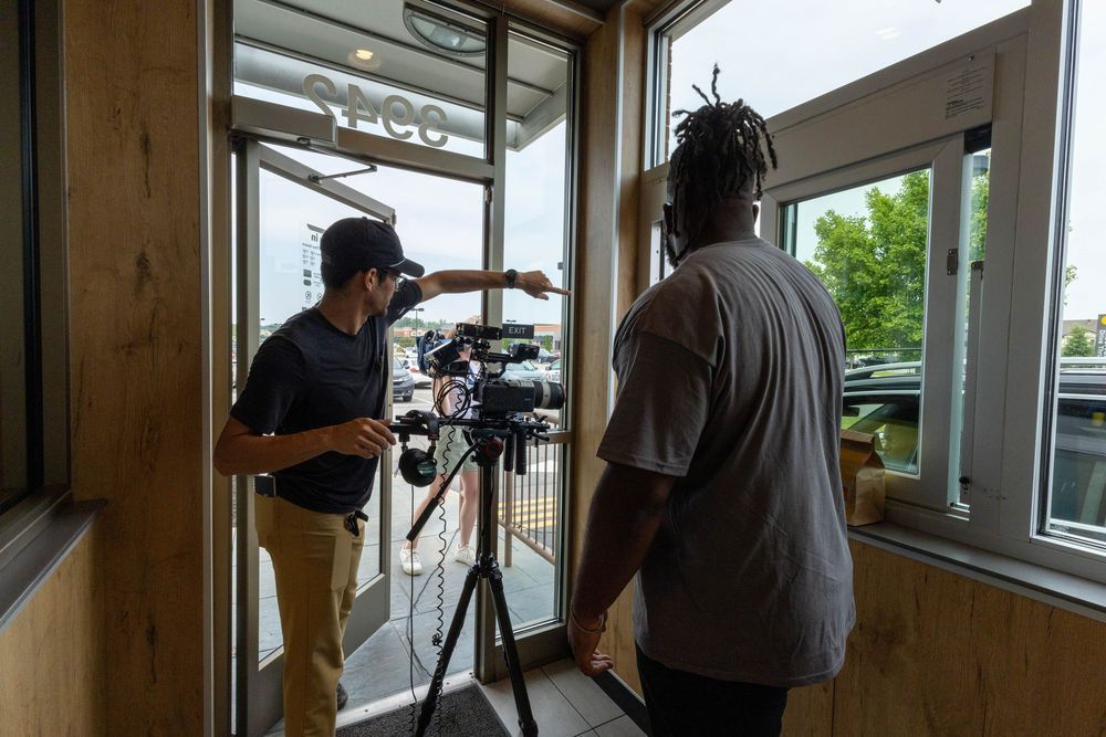 A cameraman films through a window while a man observes inside a fast food restaurant interior.