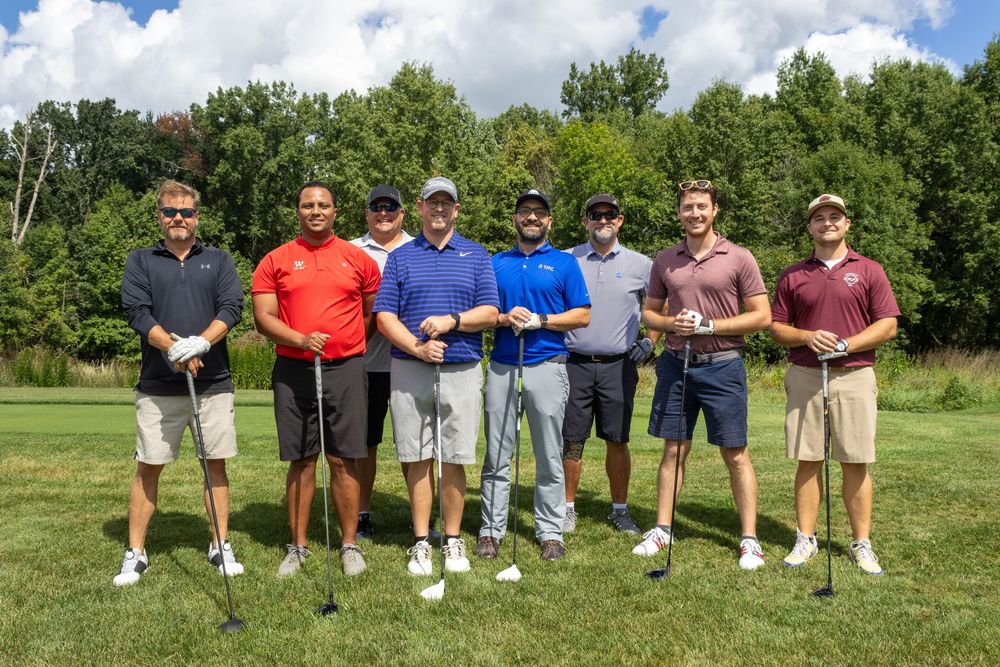 Group of seven male golfers posing together on a golf course on a sunny day.
