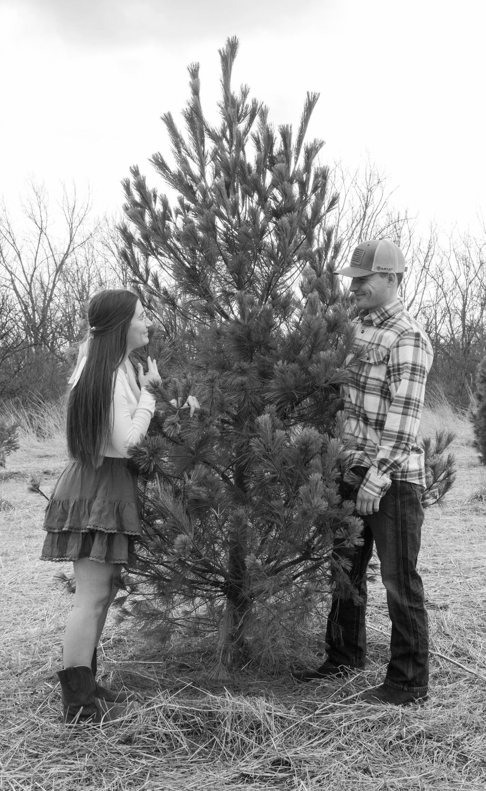 Black and white photo of young couple standing together beside a pine tree.