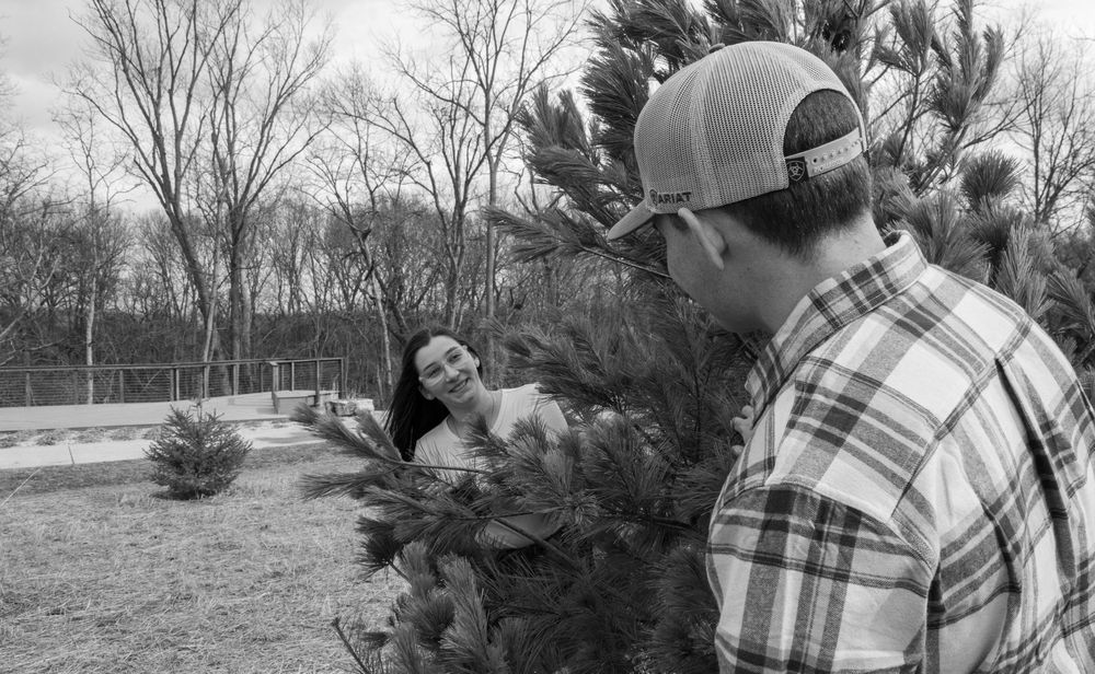 Black and white photo of couple at park behind a pine tree.