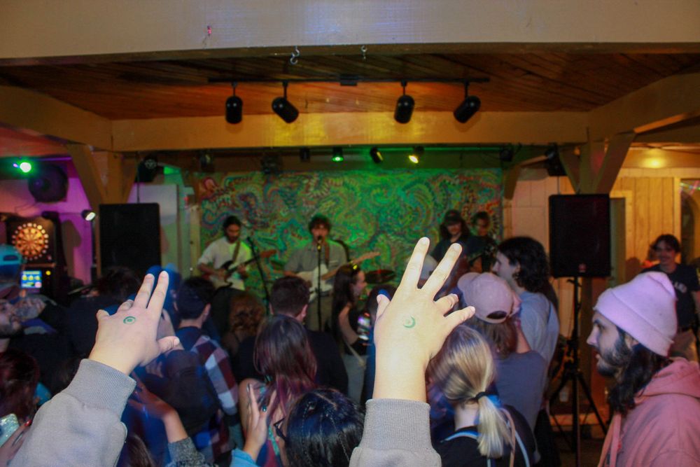 Energetic indoor concert crowd with raised hands facing a live band performing on a colorful lit stage.