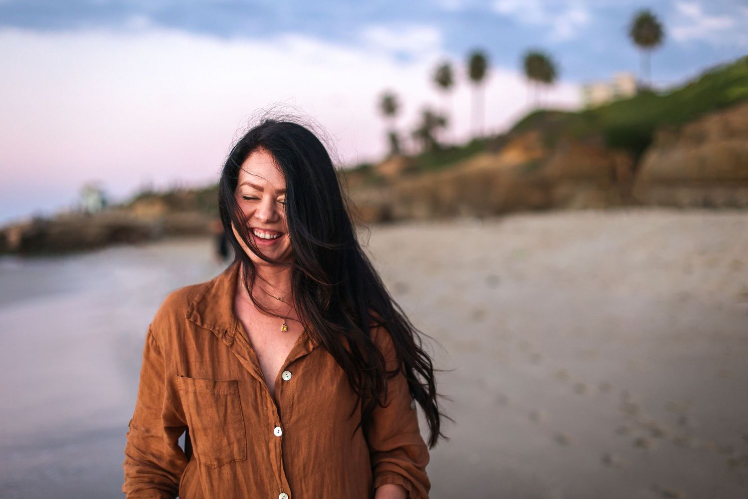 Happy Asian woman laughing on a beach at dusk with palm trees in the background.