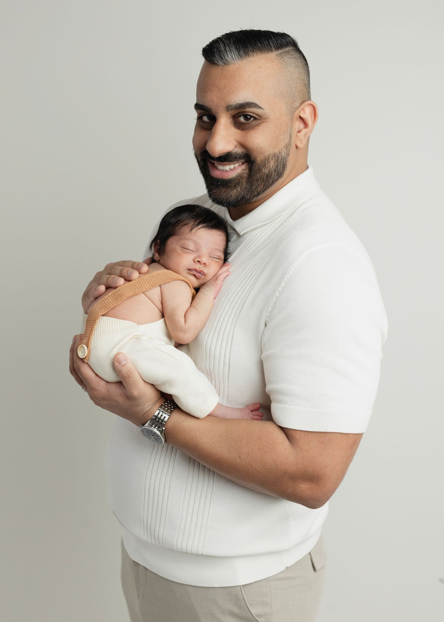 Smiling man with beard holding sleeping newborn baby wrapped in white, studio portrait photo. Elizabeth Lang Photography Surrey