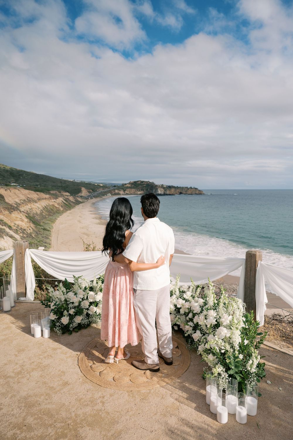 Couple embracing at a romantic beachside proposal setup with white florals and candles overlooking a scenic ocean view.