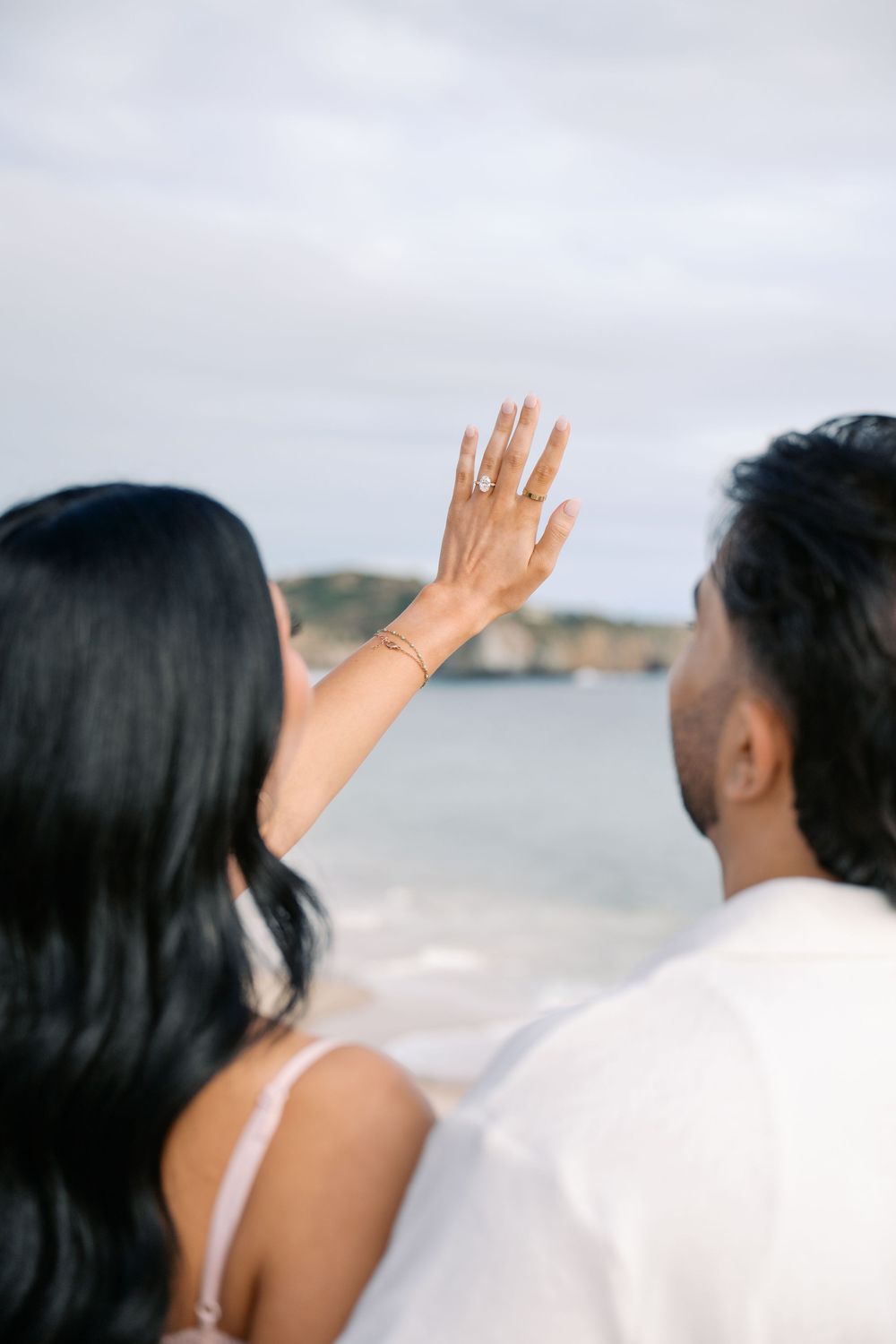 Couple at beach showing off engagement ring, woman raises hand displaying diamond ring with ocean backdrop.