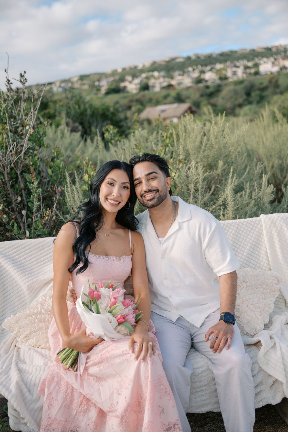 Couple sitting on white sofa outdoors, woman in pink dress holding tulip bouquet, man in white shirt, greenery background.