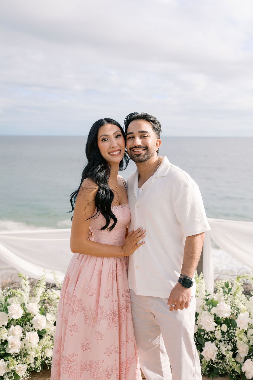 Happy couple posing together at an oceanfront proposal setup surrounded by white flowers and flowing white draping.