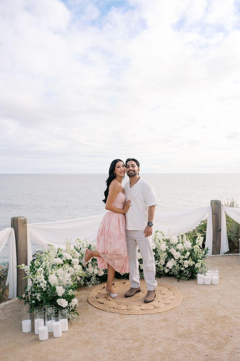 Couple embracing at cliffside ocean overlook with white floral arrangements and candles at sunset proposal setup.