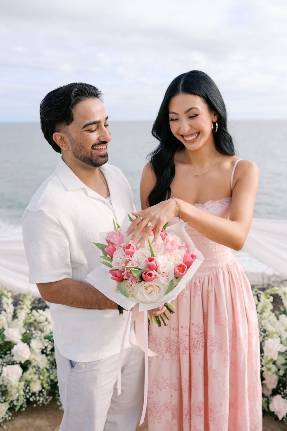 Smiling couple admiring an engagement ring while holding a pink and white tulip bouquet at a beachside proposal.