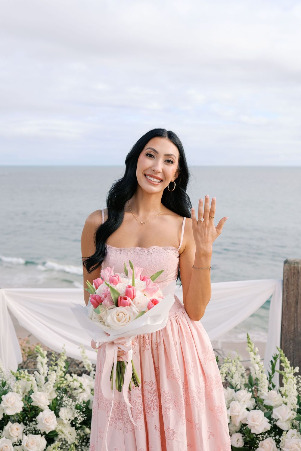 Smiling bride in pink dress shows engagement ring, holding bouquet by ocean floral arch.