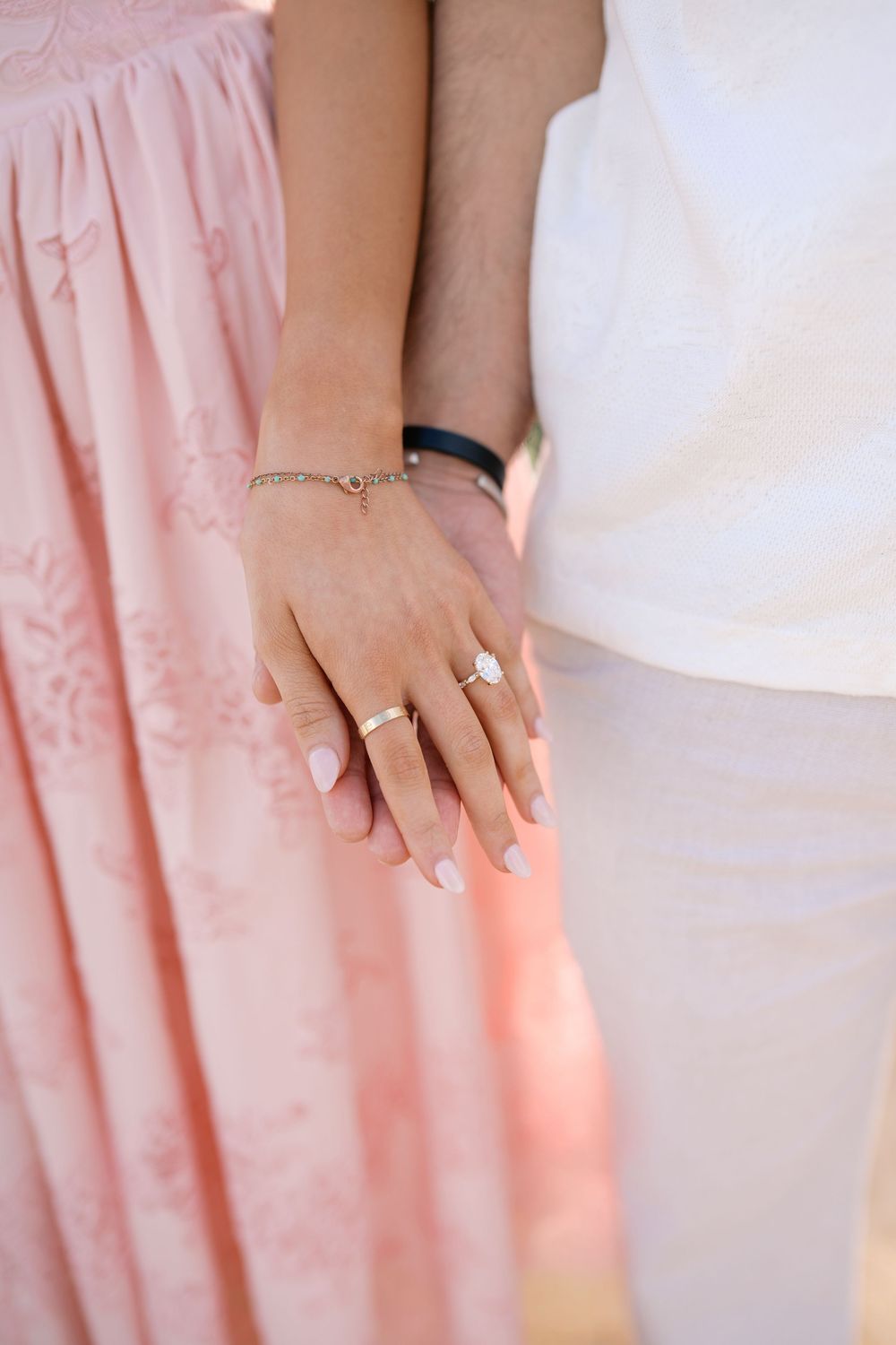 Close-up of couple holding hands, woman wearing elegant oval diamond engagement ring and delicate gold bracelet.