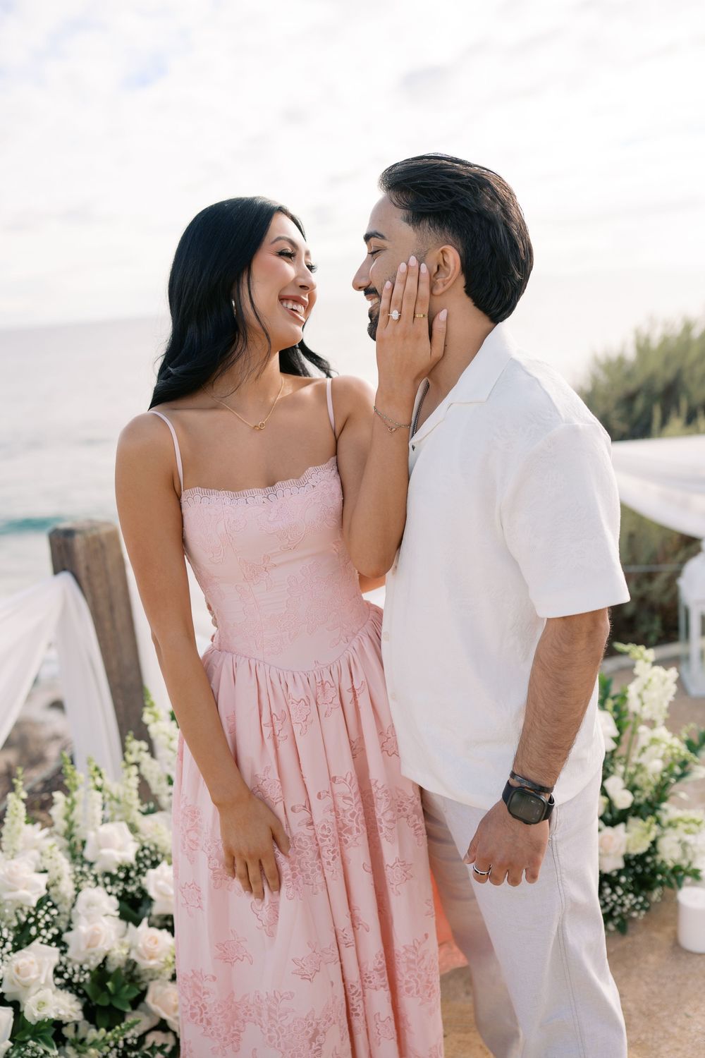 Couple smiling at each other at a romantic beach ceremony, woman in pink dress, man in white outfit with white floral decor.