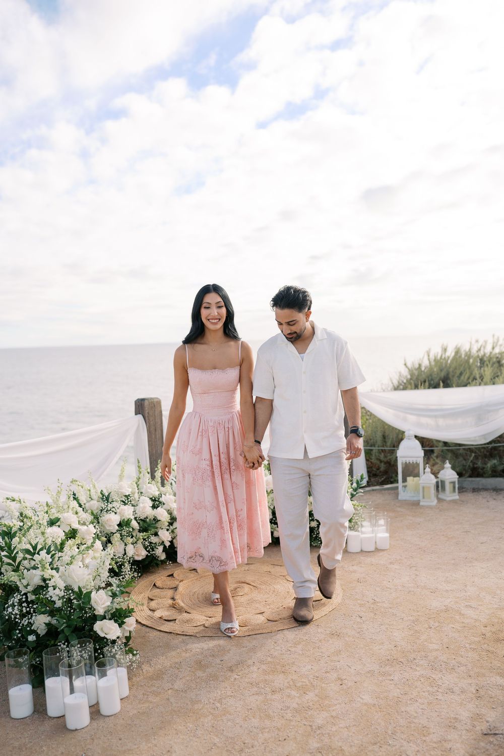 Couple holding hands at a beachside wedding ceremony with white floral arrangements and candles overlooking the ocean.