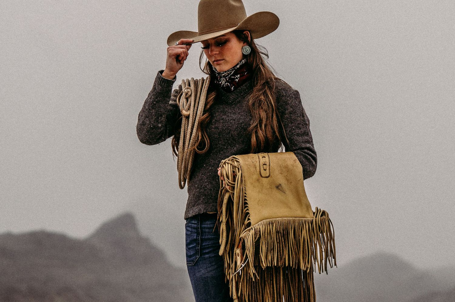Woman in cowboy hat and fringe suede crossbody bag posing outdoors with mountains in the background.