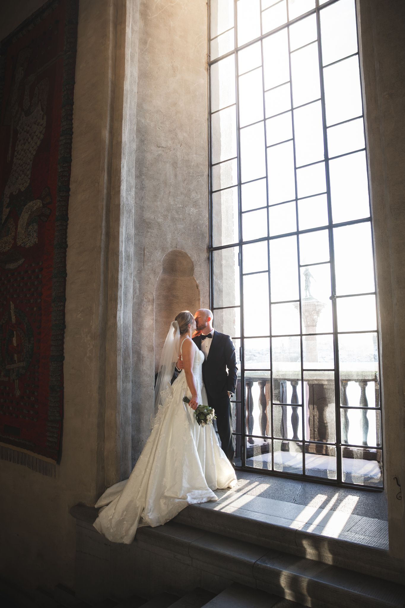 A wedding couple stands silhouetted against tall windows in a historic building, creating a romantic backlit scene.