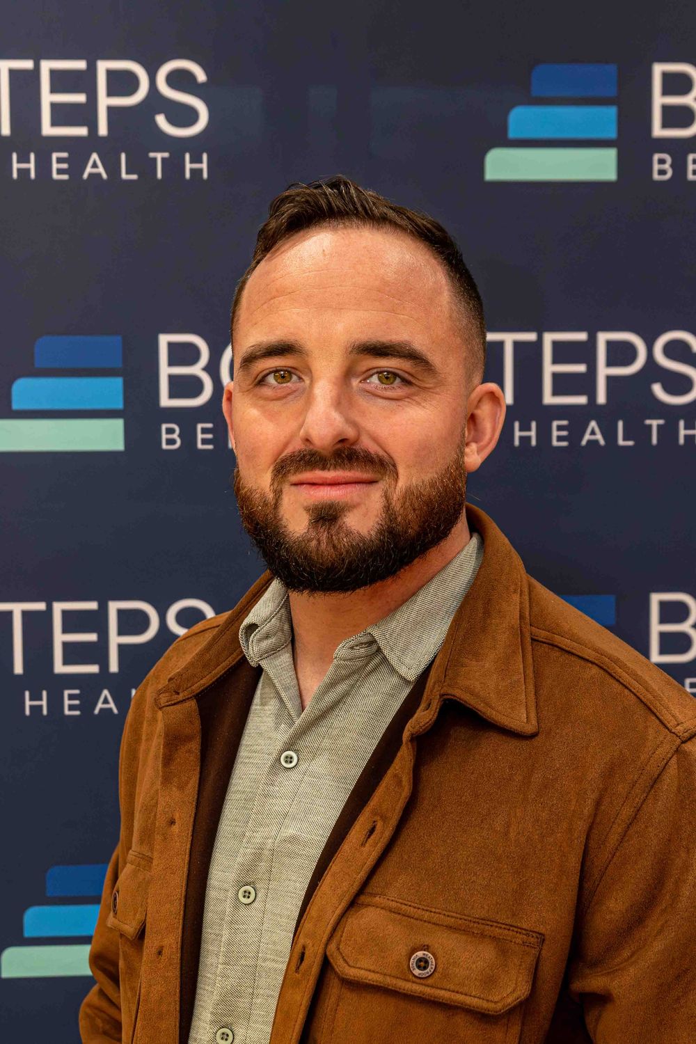 Man with beard in brown jacket smiling at Bold Steps Behavioral Health event.