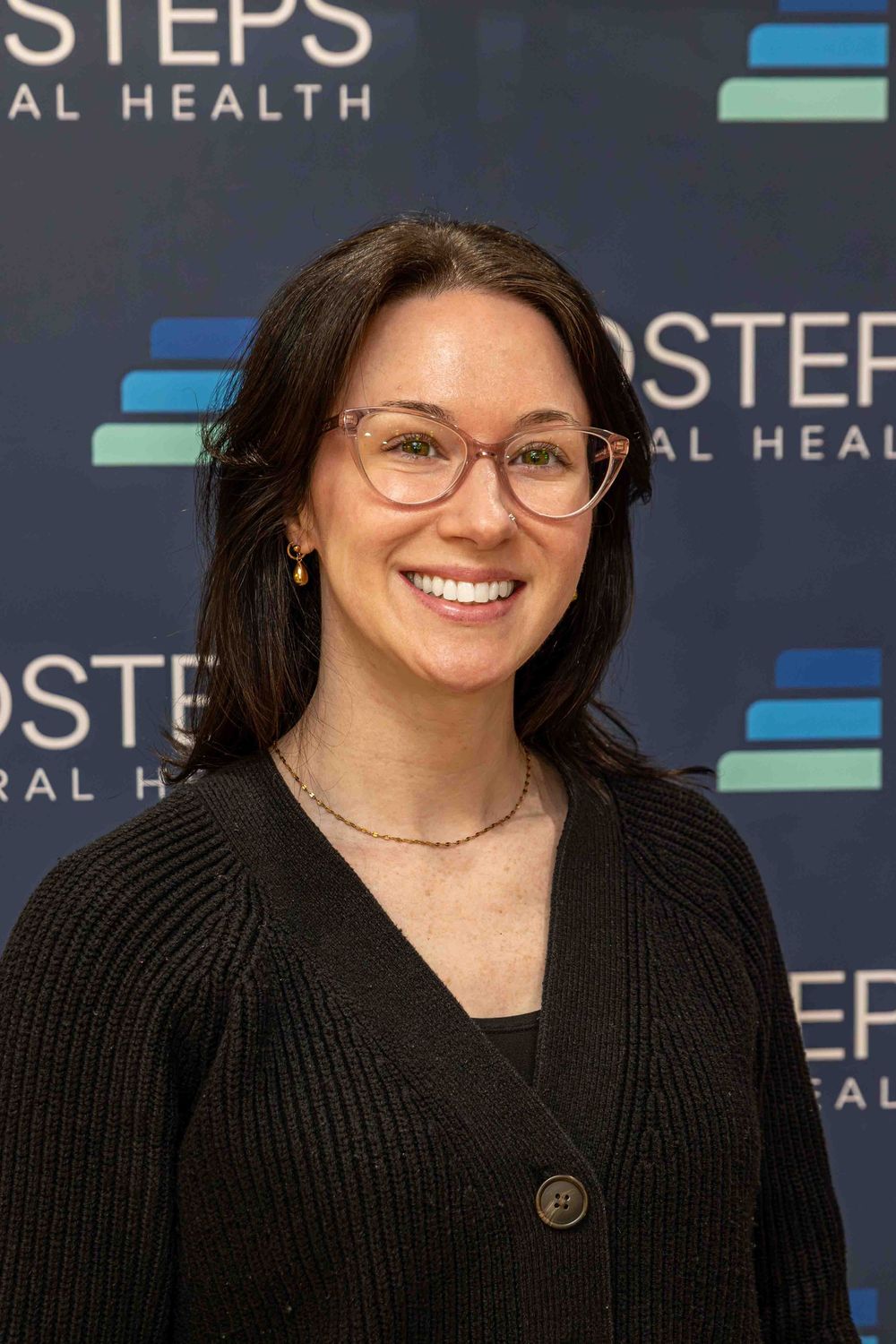 Smiling woman with glasses and dark hair poses in front of a NextSteps Behavioral Health backdrop.