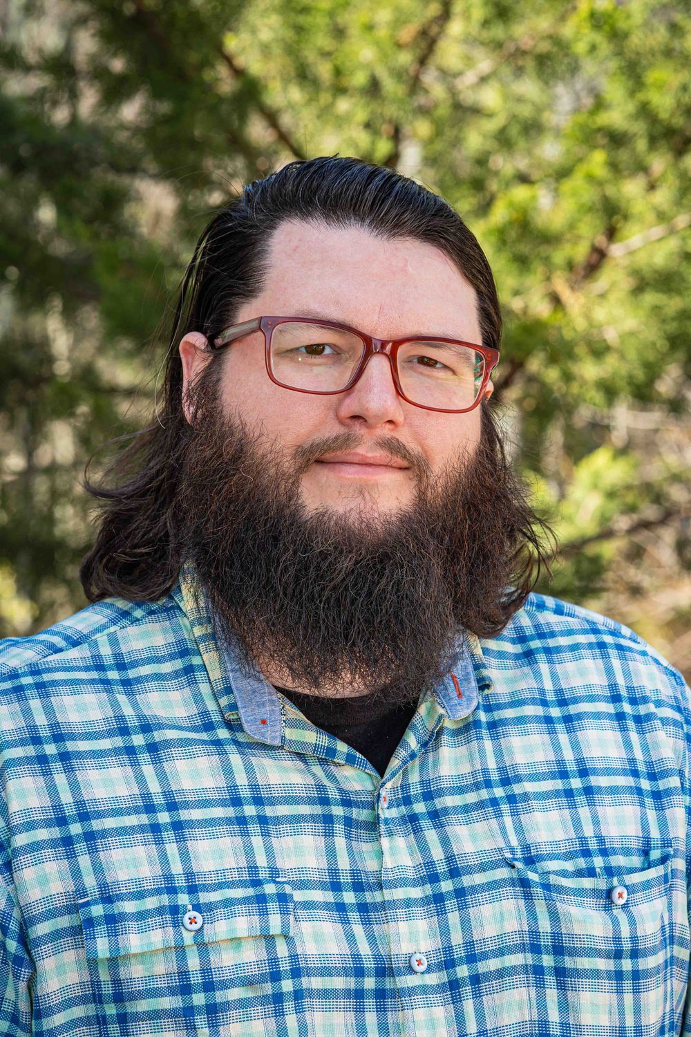 Bearded man with red glasses and long hair wearing a blue plaid shirt outdoors among green trees.