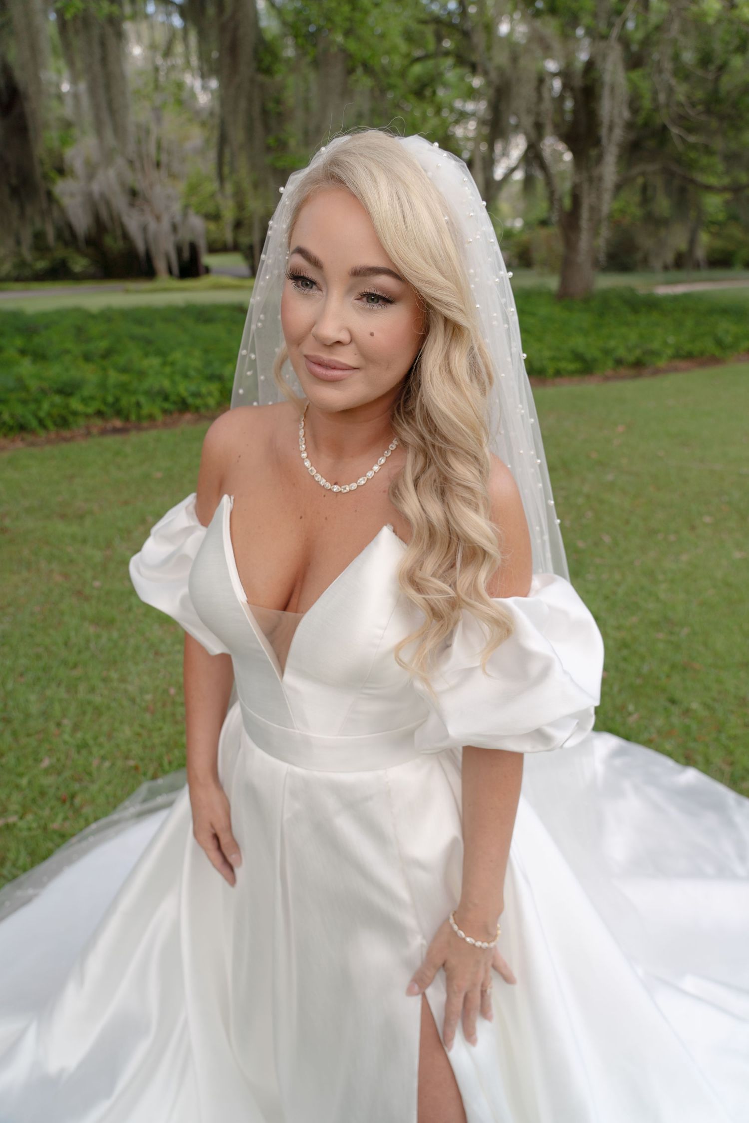 Blonde bride in white satin off-shoulder gown with veil and pearl jewelry poses outdoors among lush green trees.