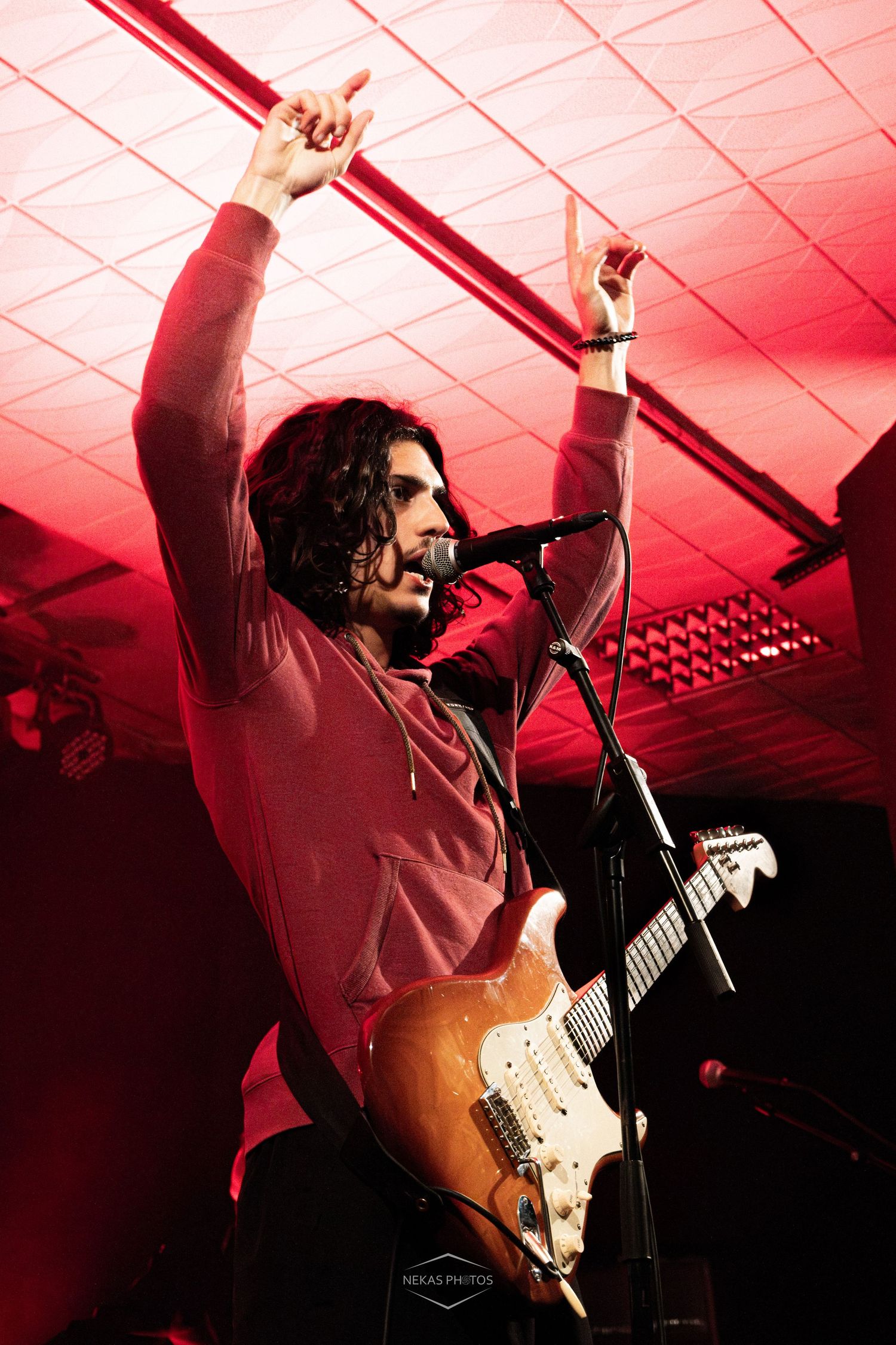 Guitarist singer performing on stage under red lights, raising hand while playing electric guitar at live concert.