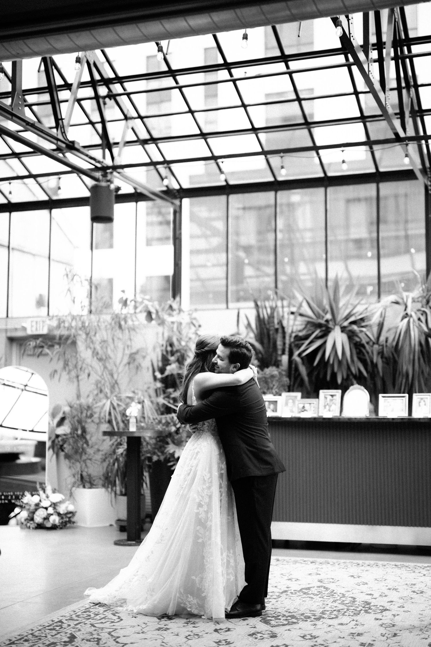 Black and white photo of bride and groom sharing first dance embrace in elegant greenhouse venue.