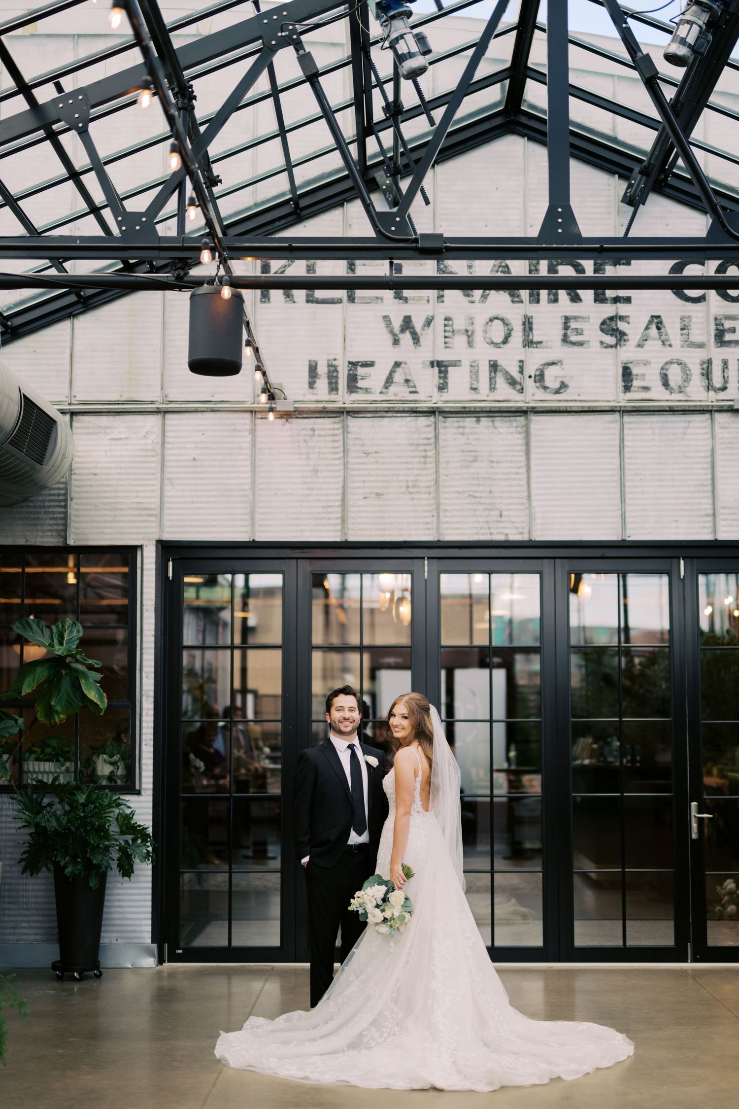 Bride and groom posing elegantly inside industrial wedding venue with glass ceiling and greenery.