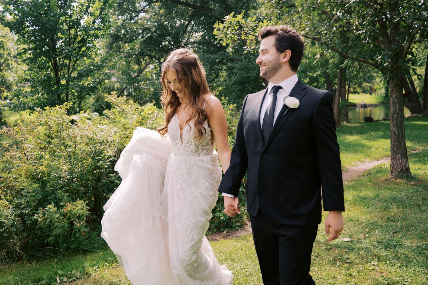 A bride and groom walk hand in hand through a lush green garden, she in a flowing white gown, he in a black suit.