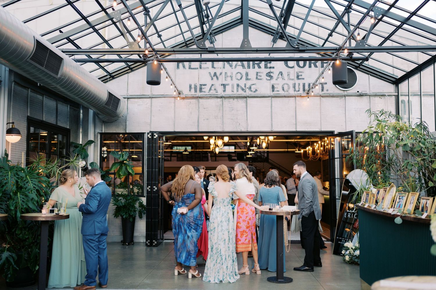 Guests mingling inside industrial greenhouse venue with lush plants during elegant wedding reception.