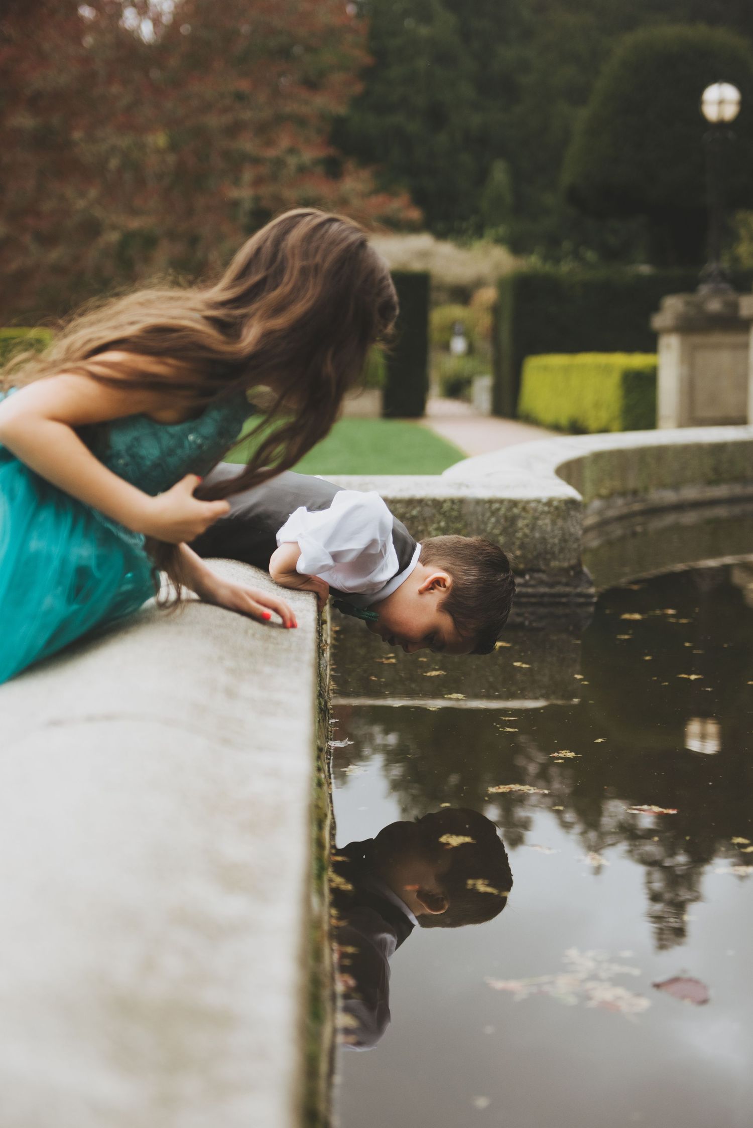 A young boy leans over the edge of a fountain to look at his reflection, while a girl in a green dress watches curiously