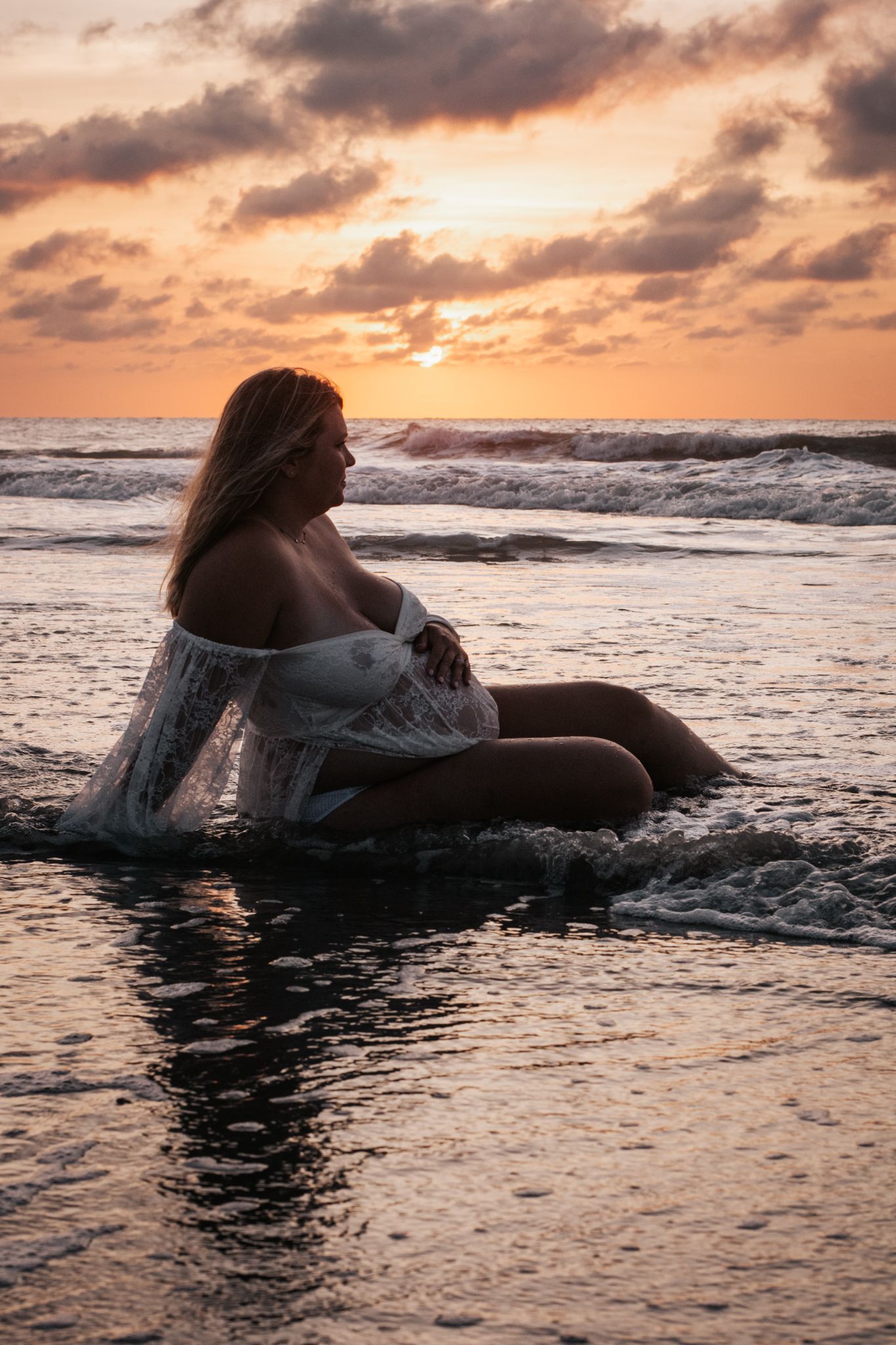A silhouette of a person sitting in shallow ocean waves during a vibrant sunset.