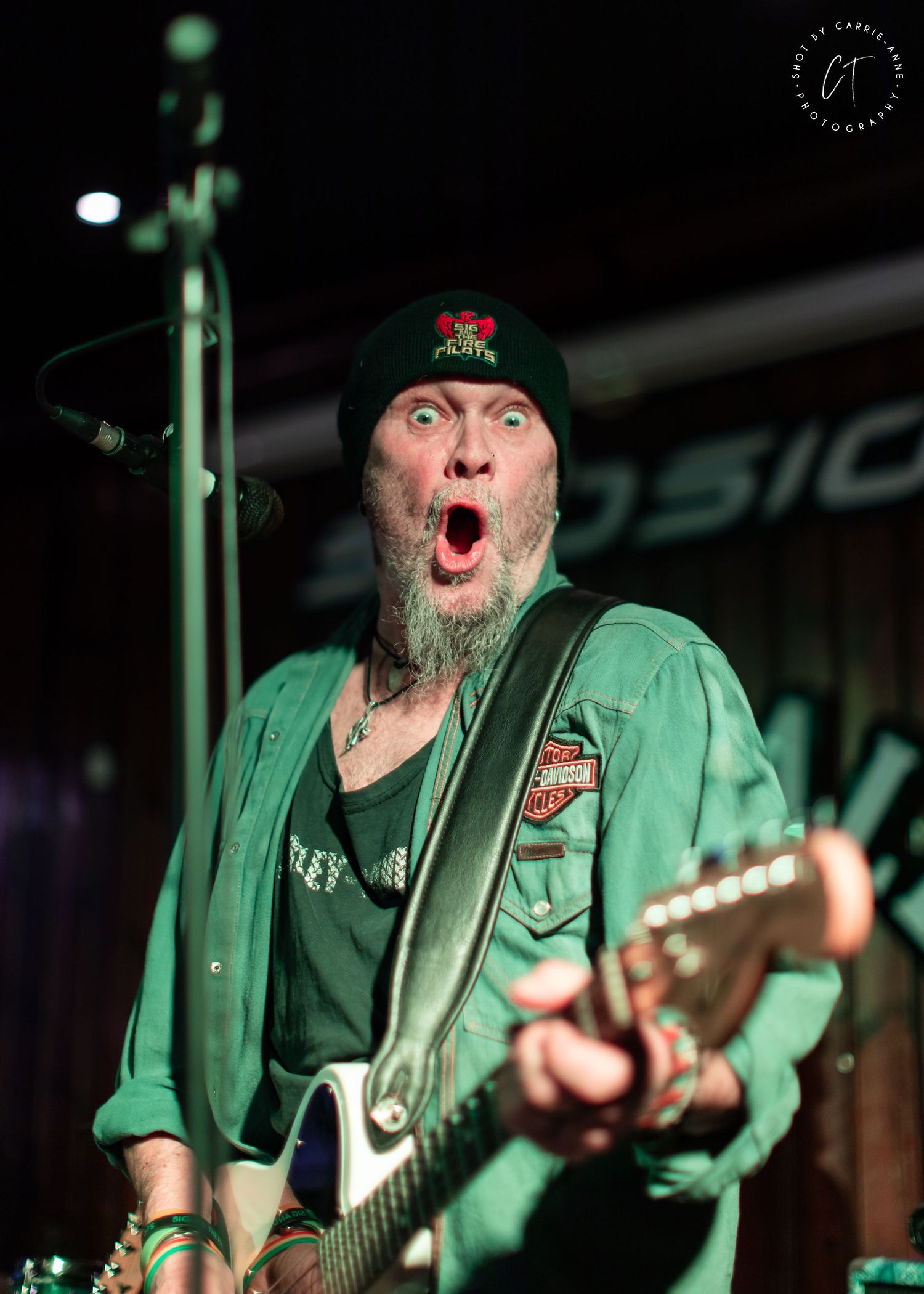 Musician in green jacket performs with guitar under colorful stage lights.