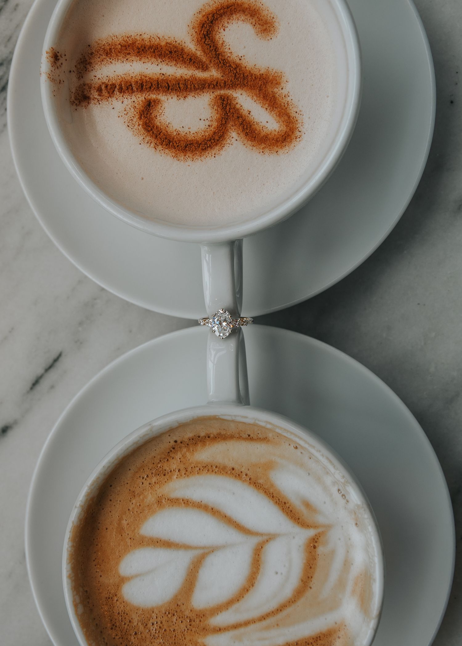 Two lattes on marble table, one with cinnamon art and one with leaf latte art, diamond ring between cups.
